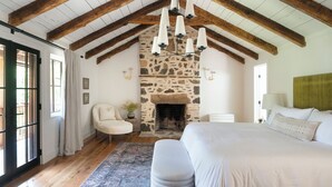 A bedroom with a stone fireplace, wooden beams, a large bed, a chair, and a rug. - The Peregrine: Flat Mountain Farm - expansive luxury home on historic 28 acre rhododendron farm (Highlands)