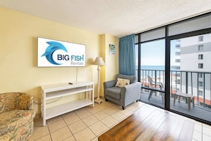 Quarto mobiliado com sofá, cadeira, mesa consola, abajur e uma janela grande com vista para um edifício e uma varanda.