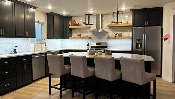 A modern kitchen with dark cabinetry, a central island with bar stools, a stainless steel refrigerator, and pendant lighting. - The Great Plains Escape (Enid)