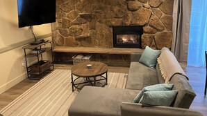 A living room with a stone wall, a fireplace, a flat-screen TV, a coffee table, and a sofa. - Mountainside ski-in condo near Ski Time Square  (Steamboat Springs)