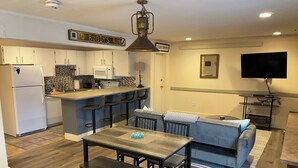 A modern kitchen with white cabinets, a microwave, and a sink. There is a bar area with stools and a sign that reads "BUDDY'S RUN". A dining table with benches is in the center, and a sofa is against the wall. A flat-screen TV is mounted on the wall. - Mountainside ski-in condo near Ski Time Square  (Steamboat Springs)