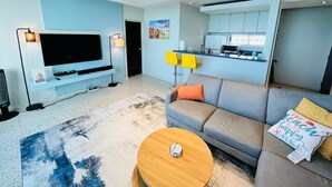 Een moderne woonkamer met een grijze bank, een salontafel, een flatscreen televisie en een keuken met lichtblauwe kastjes.