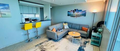 A modern living room with a grey sofa, yellow bar stools, a coffee table, and a kitchen area with a bar.