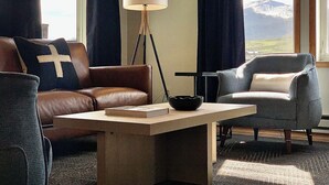Et moderne hotelværelse med en lædersofa, en grå lænestol, et lille bord med skål og bog, en gulvlampe og udsigt til bjerge gennem vinduet.