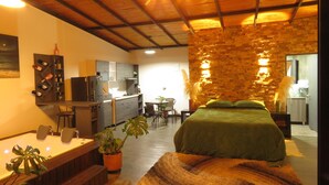 Una habitación de hotel acogedora con cama, un pequeño comedor, una cocina y una sala de estar con pared de piedra y techo de madera.