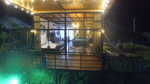 Una estructura con techo de vidrio, un deck de madera, una zona de cocina y un dormitorio visibles a través del cristal.