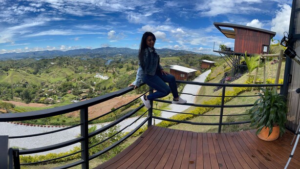 Una persona en una cubierta de madera contemplando un paisaje pintoresco con colinas, un río y un edificio moderno.