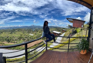 A person on a wooden deck overlooking a scenic landscape with hills, a river, and a modern building. - Hermosa Cabaña en Guatapé con Jacuzzi - Hermosa Visa y Paisaje (Guatape)
