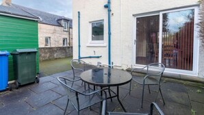Une terrasse avec une table ronde et quatre chaises, un abri de jardin vert et une maison à l’extérieur blanche avec des tuyaux bleus.