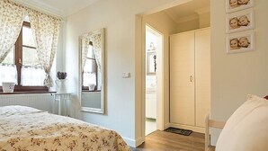 Un dormitorio con cama, ventana con cortinas, espejo, armario y puerta que da a otra habitación.