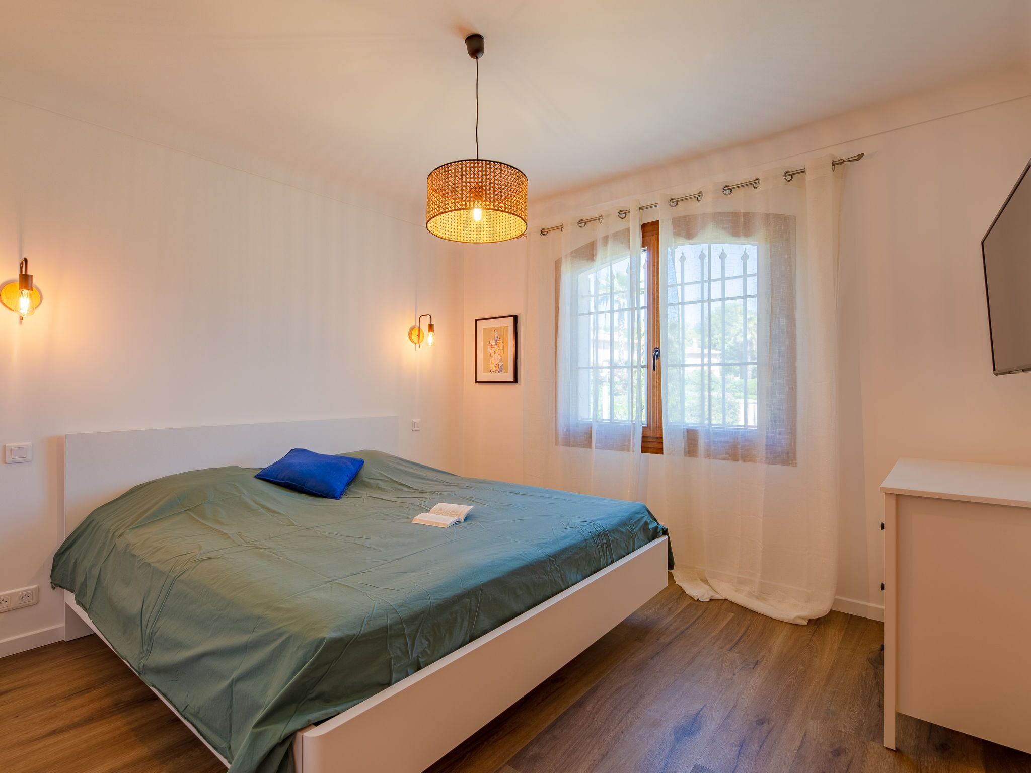 Ein Schlafzimmer mit Bett, Nachttisch, einem Fenster mit Vorhängen und einer Hängelampe.