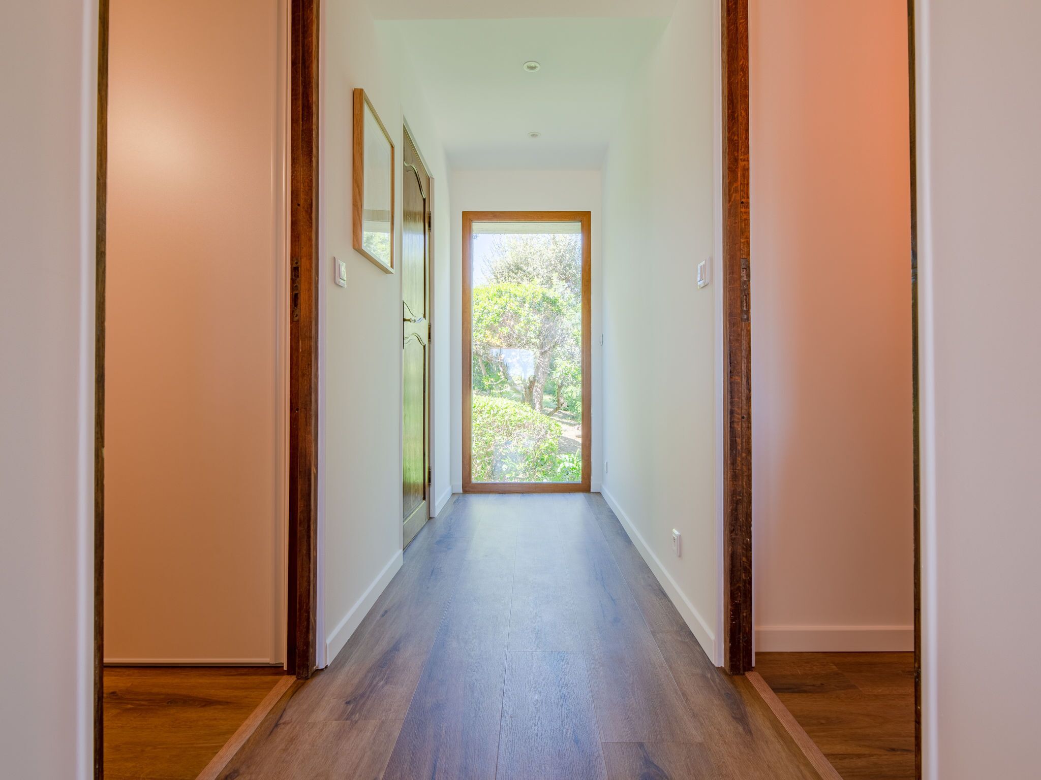A hallway with wooden flooring and white walls, leading to a glass door with a view of greenery outside.
