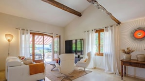 Een ruime woonkamer met een witte bank, twee witte fauteuils, een houten salontafel en een televisie. Grote ramen met gordijnen laten natuurlijk licht binnen, en een houten balk in het plafond zorgt voor een rustieke sfeer.