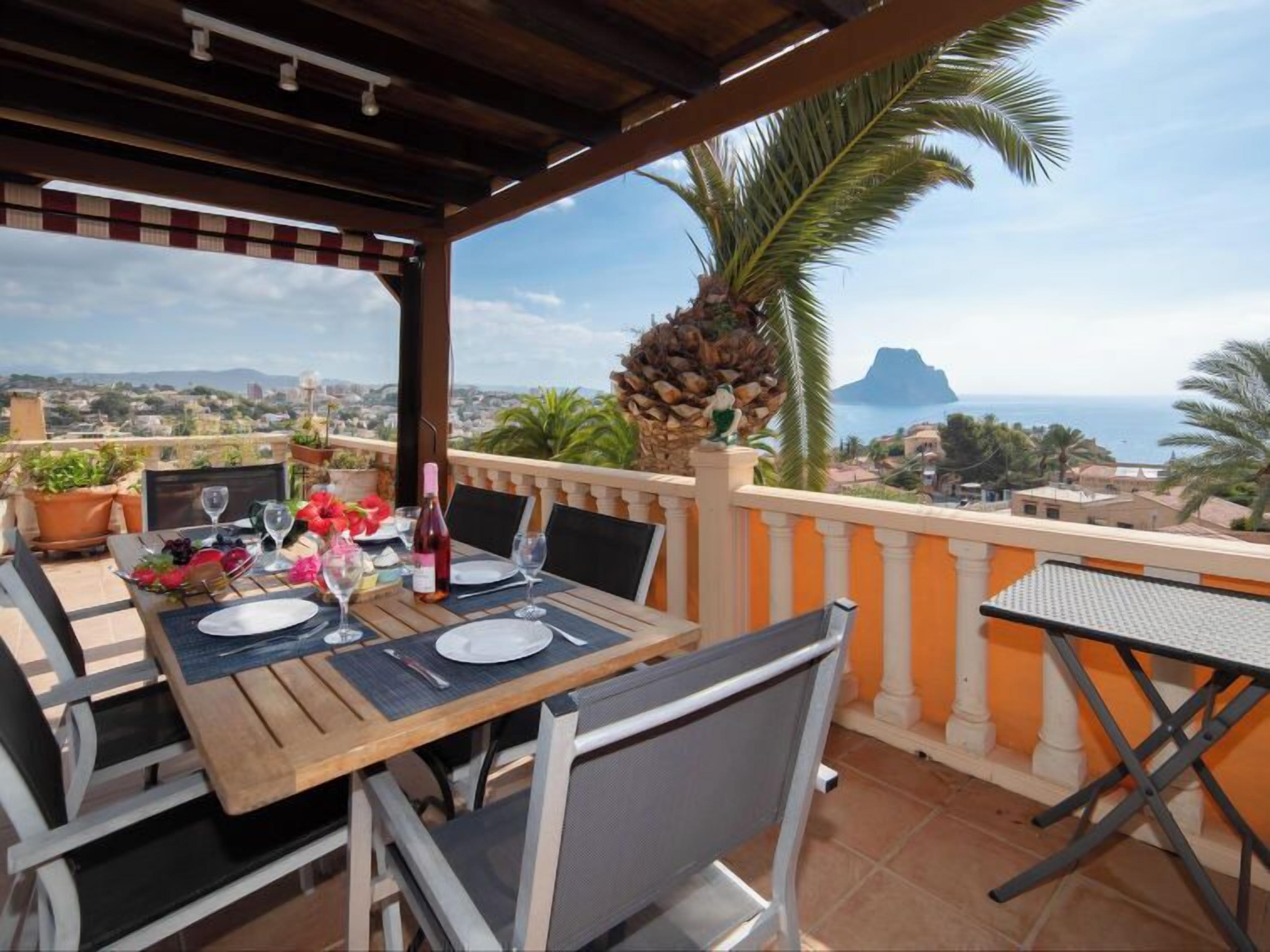 Eine Terrasse mit Holztisch, gedeckt für ein Essen, mit Blick auf die Küste, Palmen und einen entfernten Berg.