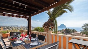 Eine Terrasse mit Holztisch, gedeckt für ein Essen, mit Blick auf die Küste, Palmen und einen entfernten Berg.