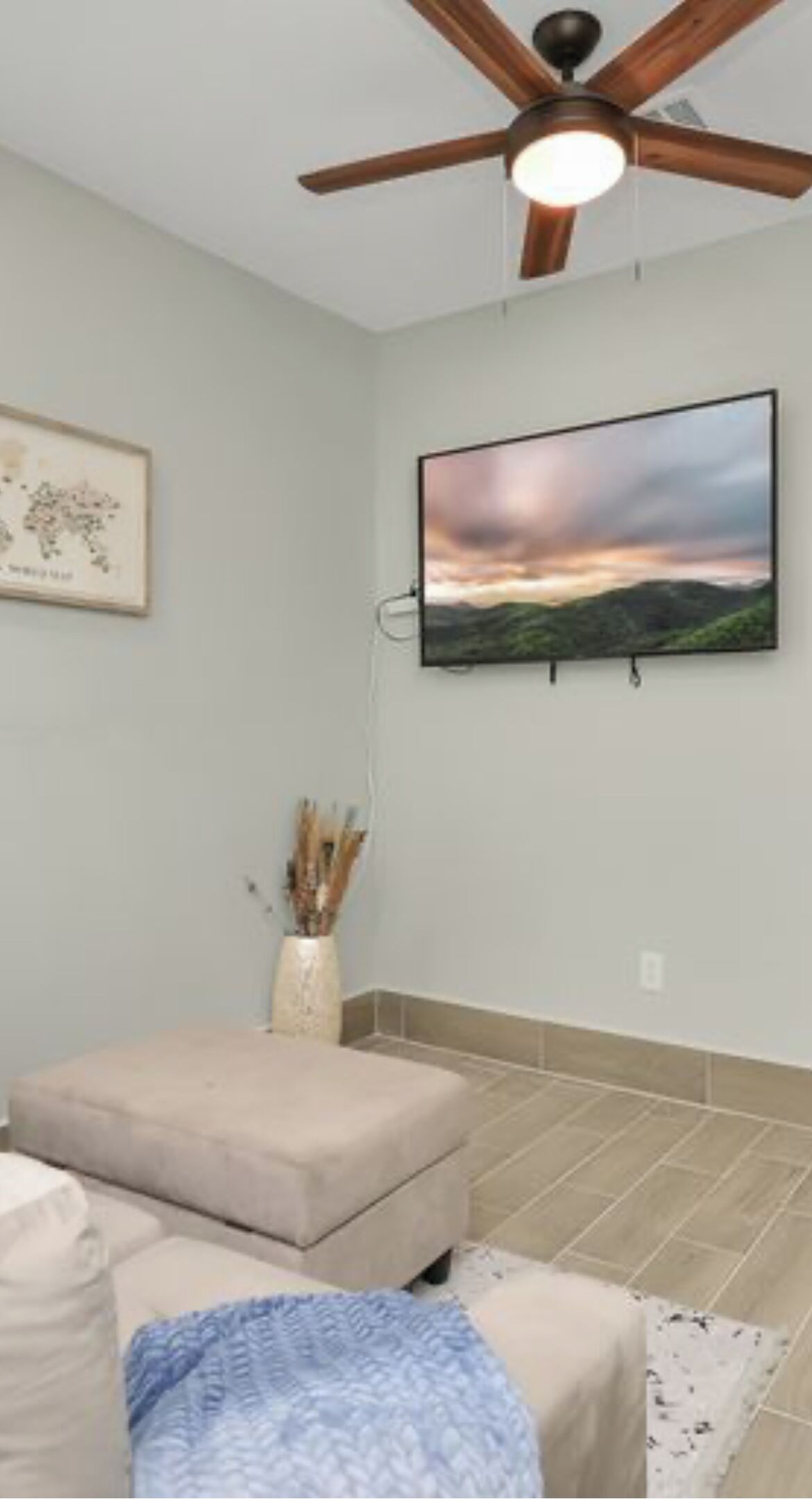 Una habitación con ventilador de techo, televisor montado mostrando un paisaje, un otoman beige y una mesita con un jarrón de plantas secas.