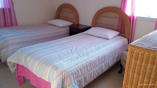 A room with two single beds, each with a wicker headboard and a striped bedspread. There is a small wooden nightstand between the beds.