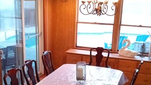 Un coin repas avec une table recouverte de plastique, entourée de chaises en bois, d’un lustre et offrant une vue sur l’extérieur par une fenêtre.