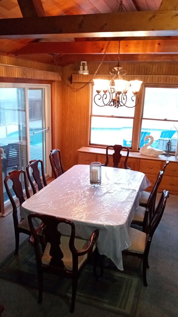 Un coin repas avec une table recouverte de plastique, entourée de chaises en bois, d’un lustre et offrant une vue sur l’extérieur par une fenêtre.