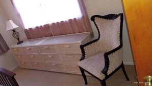 A room with a large chest of drawers, a chair, a lamp, and a window with curtains.