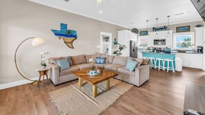 A modern living room with a wooden coffee table, beige sofa, and a kitchen area with white cabinets and a blue island. - Casa Crystal - Relax and create unforgettable memories (Bolivar Peninsula)