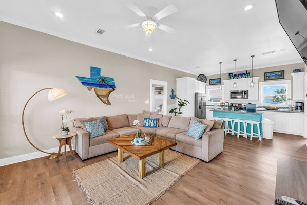 A modern living room with a wooden coffee table, beige sofa, and a kitchen area with white cabinets and a blue island.