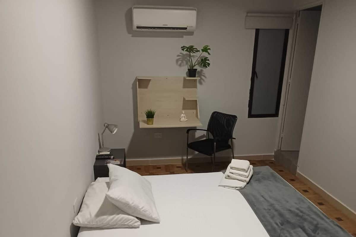A hotel room with a bed, a desk with a chair, a small plant, and a window.
