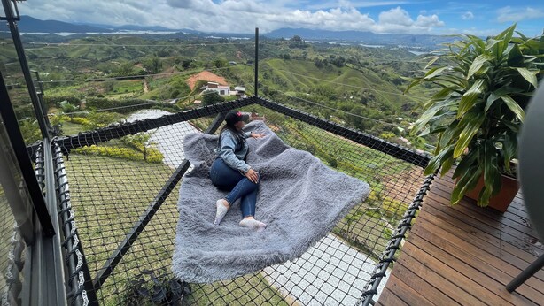 Una persona sentada sobre una manta suave de color gris dentro de una estructura con mosquitero, con vistas a un valle exuberante y colinas ondulantes, y una casa a lo lejos.