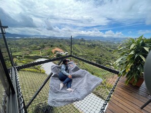 A person sitting on a soft, grey blanket inside a netted enclosure, overlooking a lush valley with rolling hills and a distant house.