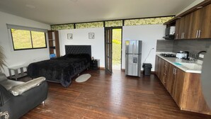Un dormitorio moderno con una cama grande, un sofá pequeño y una zona de descanso separada. La habitación tiene piso de madera, un armario empotrado y una ventana con vistas a la vegetación.