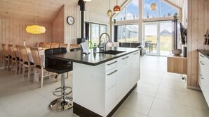 A modern kitchen with a central island, large windows, and a dining area with wooden chairs.