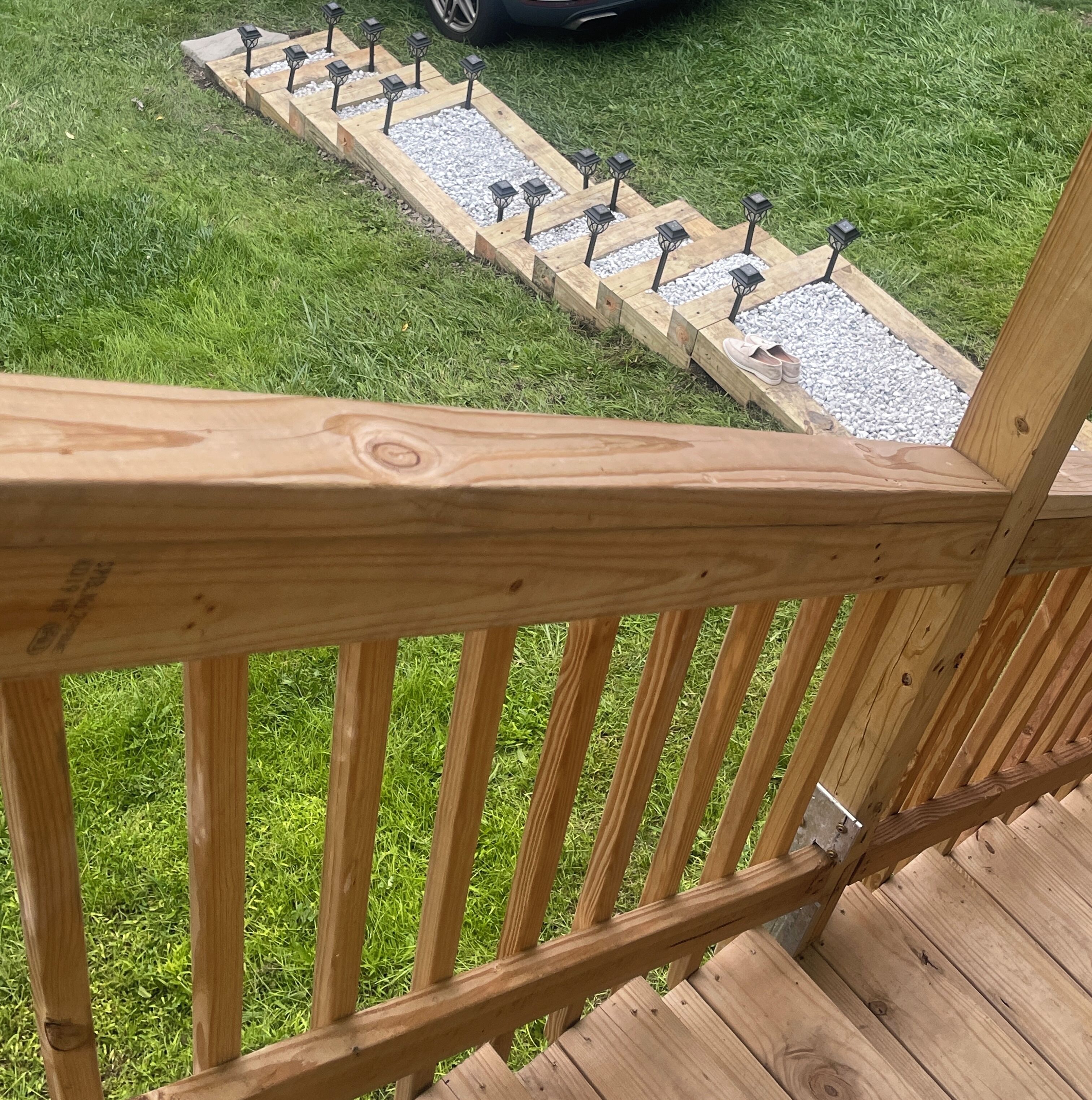 A wooden deck with a railing, a pathway with lanterns, and a car in the background.
