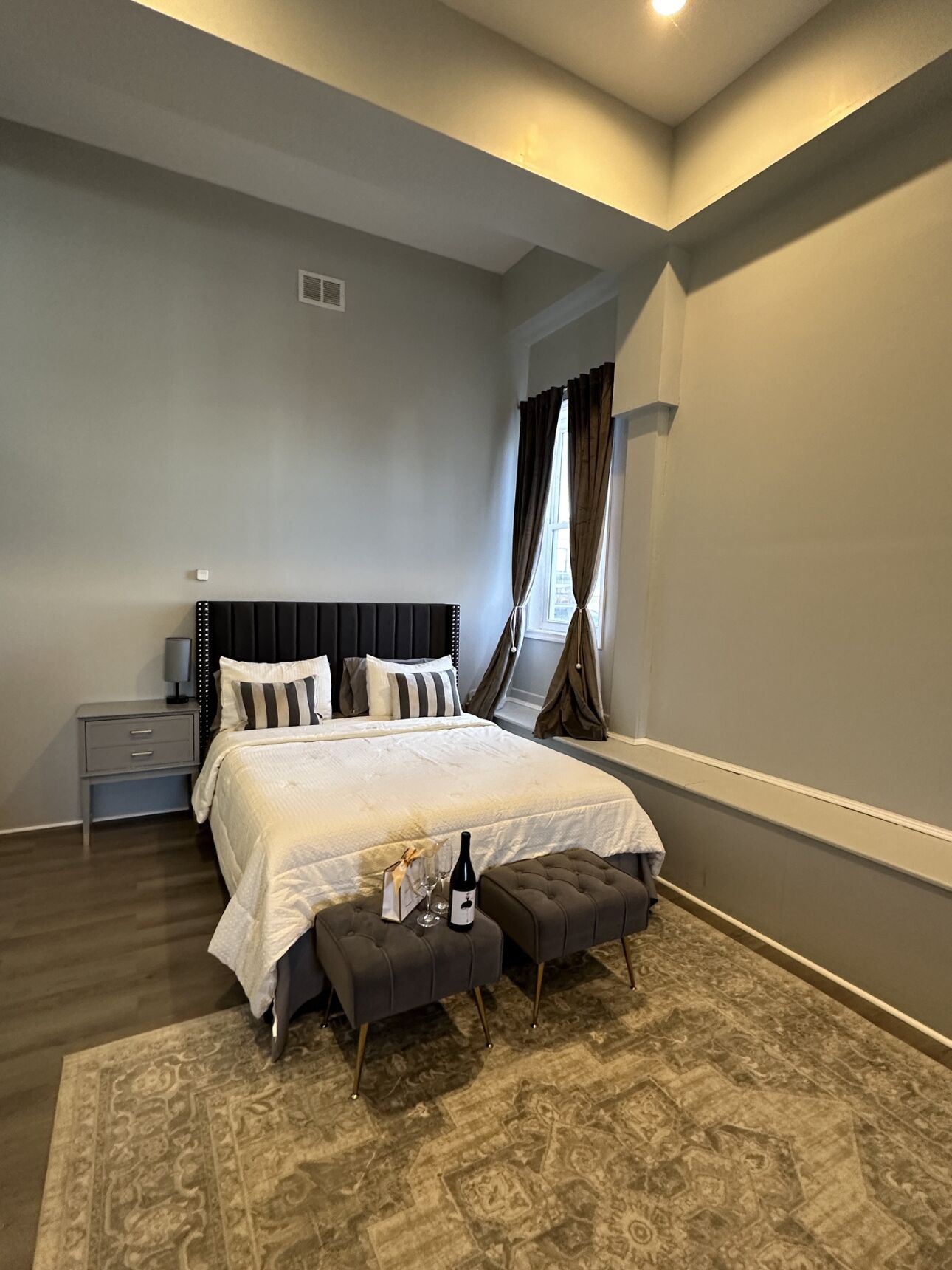 A modern bedroom with a large bed, a nightstand, a bedside table, a window with curtains, and a patterned rug.