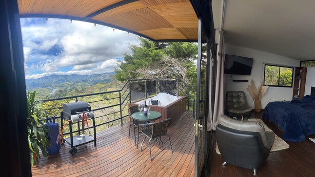 A balcony with a wooden deck, a barbecue grill, a table with chairs, and a view of the landscape.