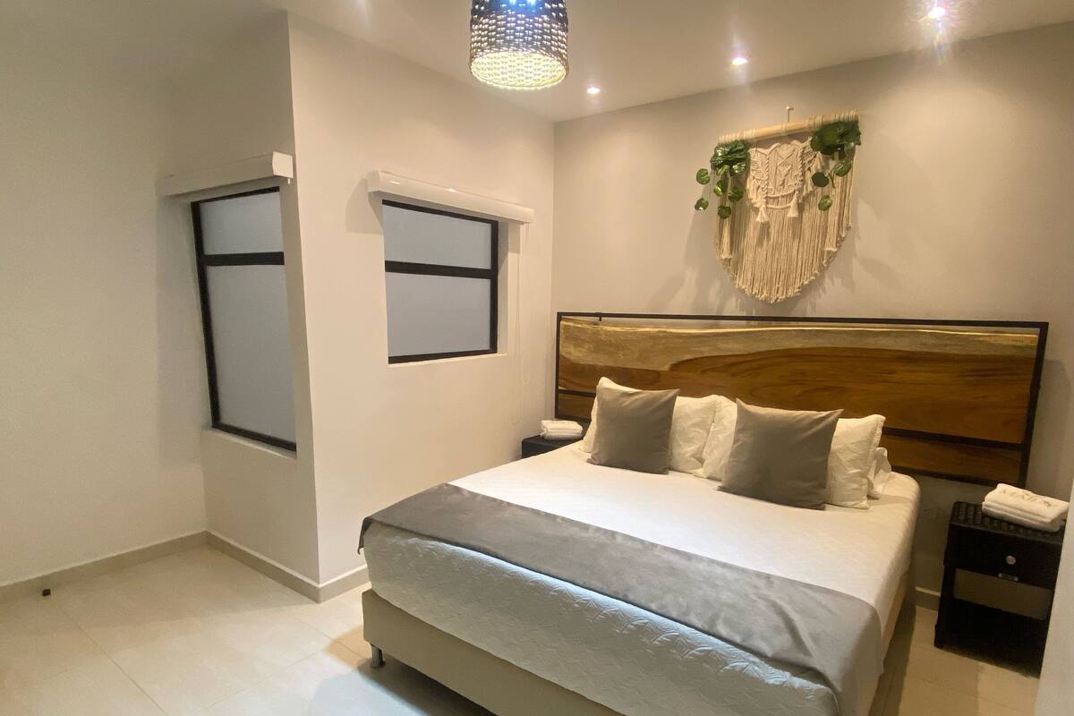Une chambre moderne avec un grand lit, une table de chevet, une décoration murale en macramé et une fenêtre avec des stores.
