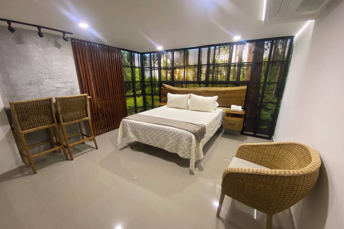 Une chambre à coucher comprenant un lit, deux fauteuils en osier, une table de chevet et une grande fenêtre donnant sur des arbres.