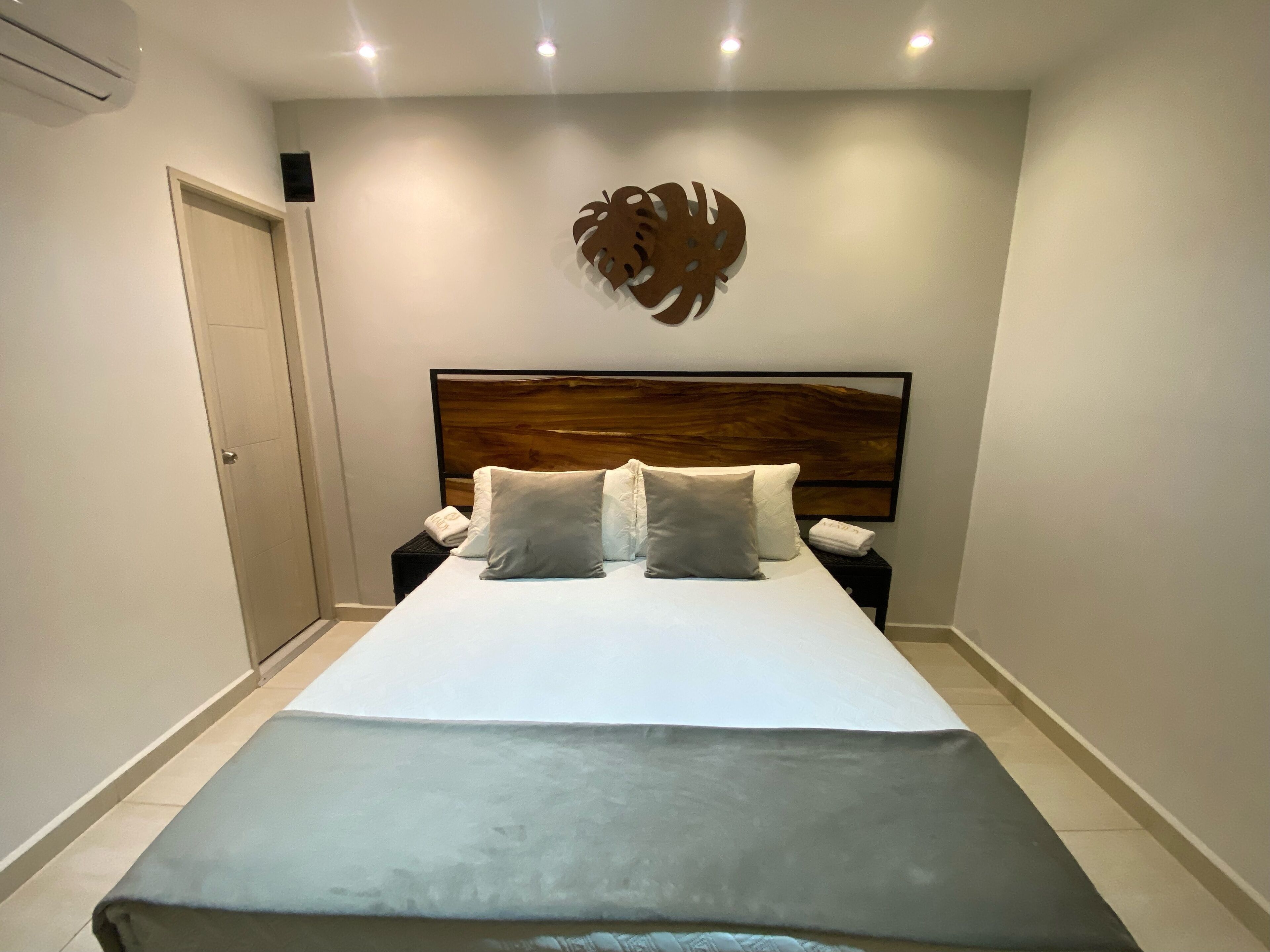 A neatly made bed with a grey bedspread and pillows, a wooden headboard, a wall-mounted decorative piece, and a nightstand with a phone and a folded towel.