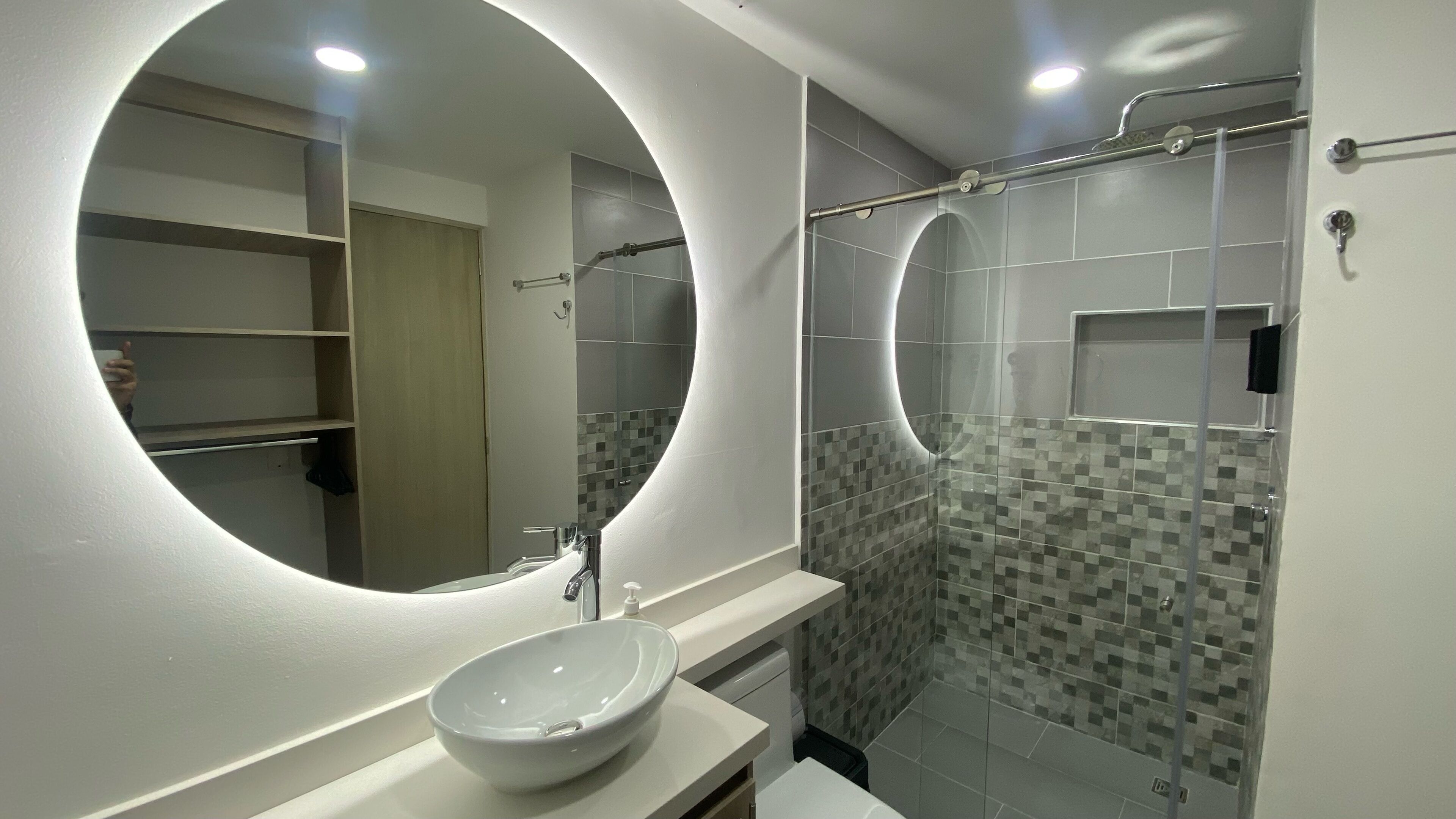 Une salle de bain moderne avec un miroir rond, un lavabo blanc et une douche dotée d’une porte en verre.