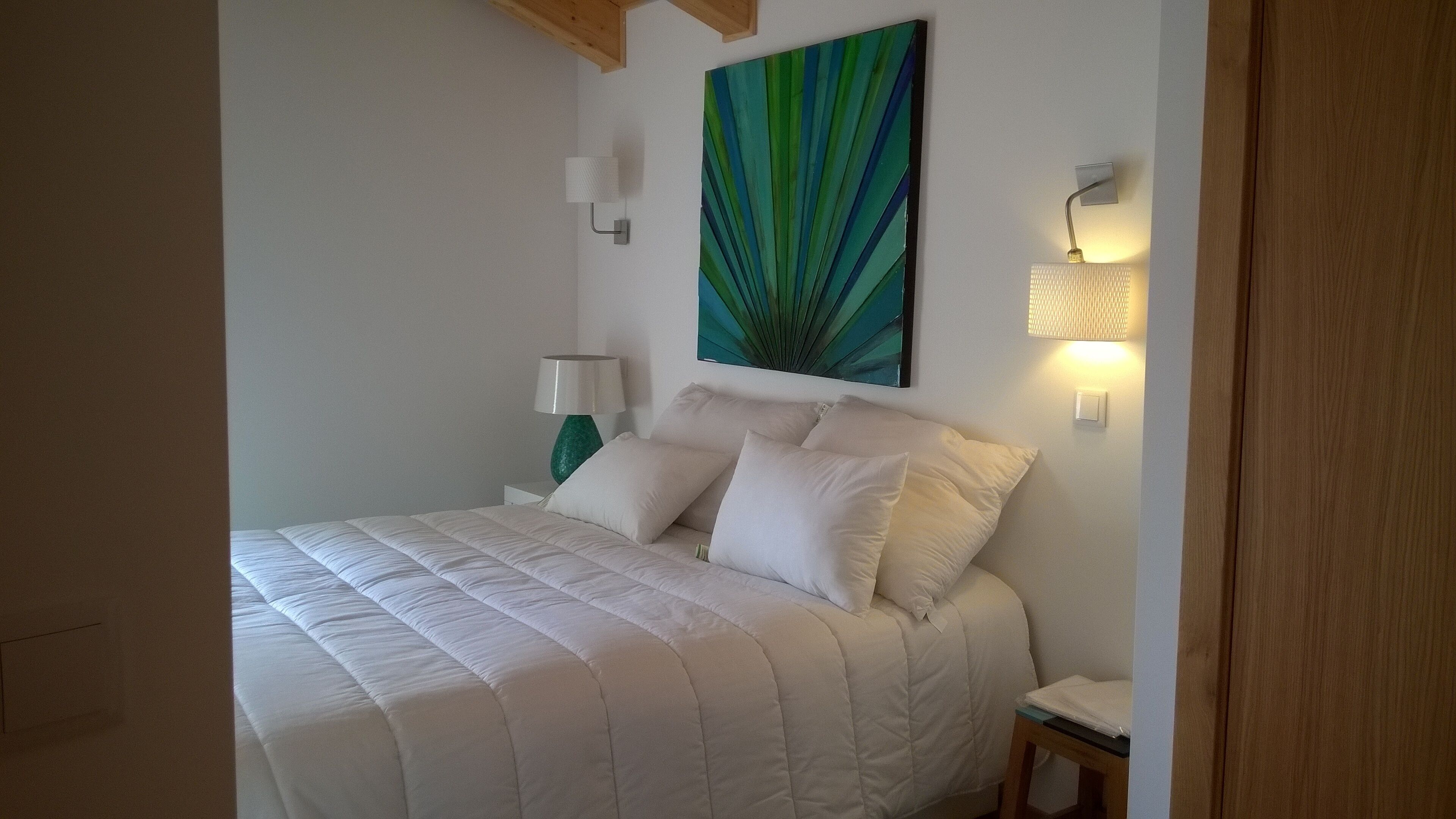 Una cama bien hecha con ropa de cama y almohadas blancas, un cuadro abstracto verde en la pared y dos lámparas fijadas a la pared.