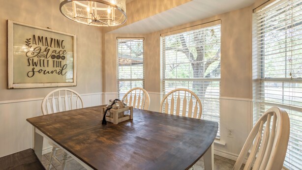 Un coin repas avec une table en bois, des chaises blanches, un lustre et une fenêtre avec des stores.