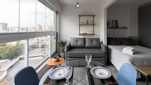 Et moderne hotelværelse med udsigt over byen, en sofa, en seng, et spisebord med tallerkener og en lille plante.