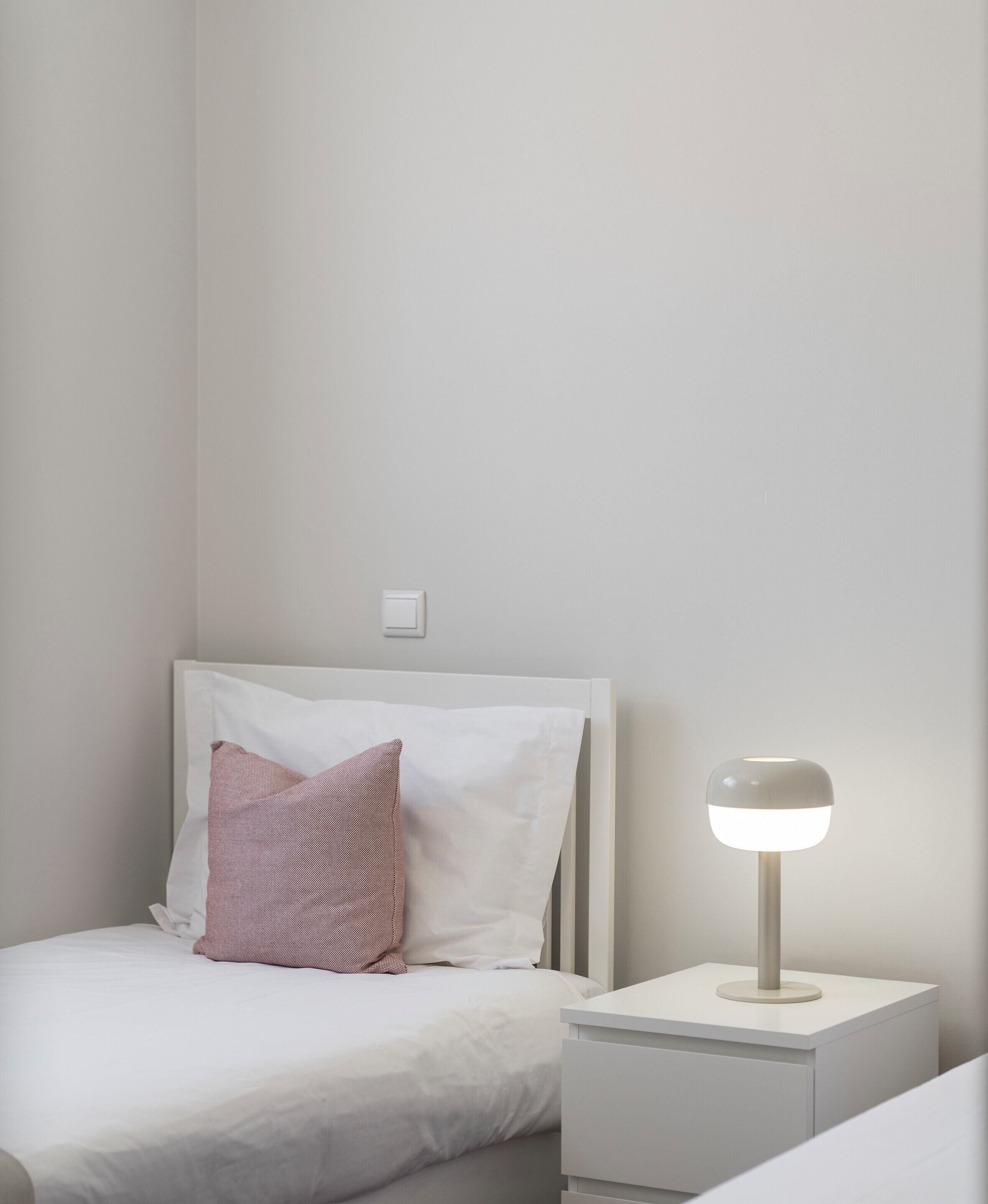 A neatly made bed with white bedding and a pink pillow, a bedside table with a lamp, and a wall switch.