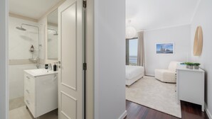 Ein modernes Hotelzimmer mit Bad, das eine Dusche, ein Waschbecken und einen Spiegel beinhaltet. Das Zimmer verfügt über ein Bett, einen kleinen Nachttisch mit Pflanze und Meerblick durch ein großes Fenster.