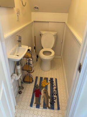 A small bathroom with a white toilet, a sink, and a shower. There is a rug with cartoon characters on the floor. - Lakefront retreat w/ deck, dock, sandy beach, 4 watercraft, <6 mins to 2 beaches (Orleans)