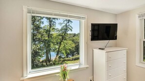 Huoneessa on suuri ikkuna, josta avautuu näkymä metsäiselle alueelle, valkoinen lipasto ja seinälle asennettu televisio.