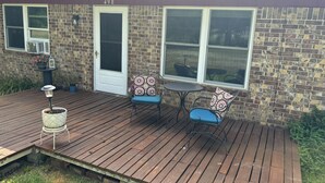 A wooden deck with two chairs and a table, a door, and a brick wall with windows. - Great get away to a country home with large yard and outdoor sitting area.  (Rose Bud)