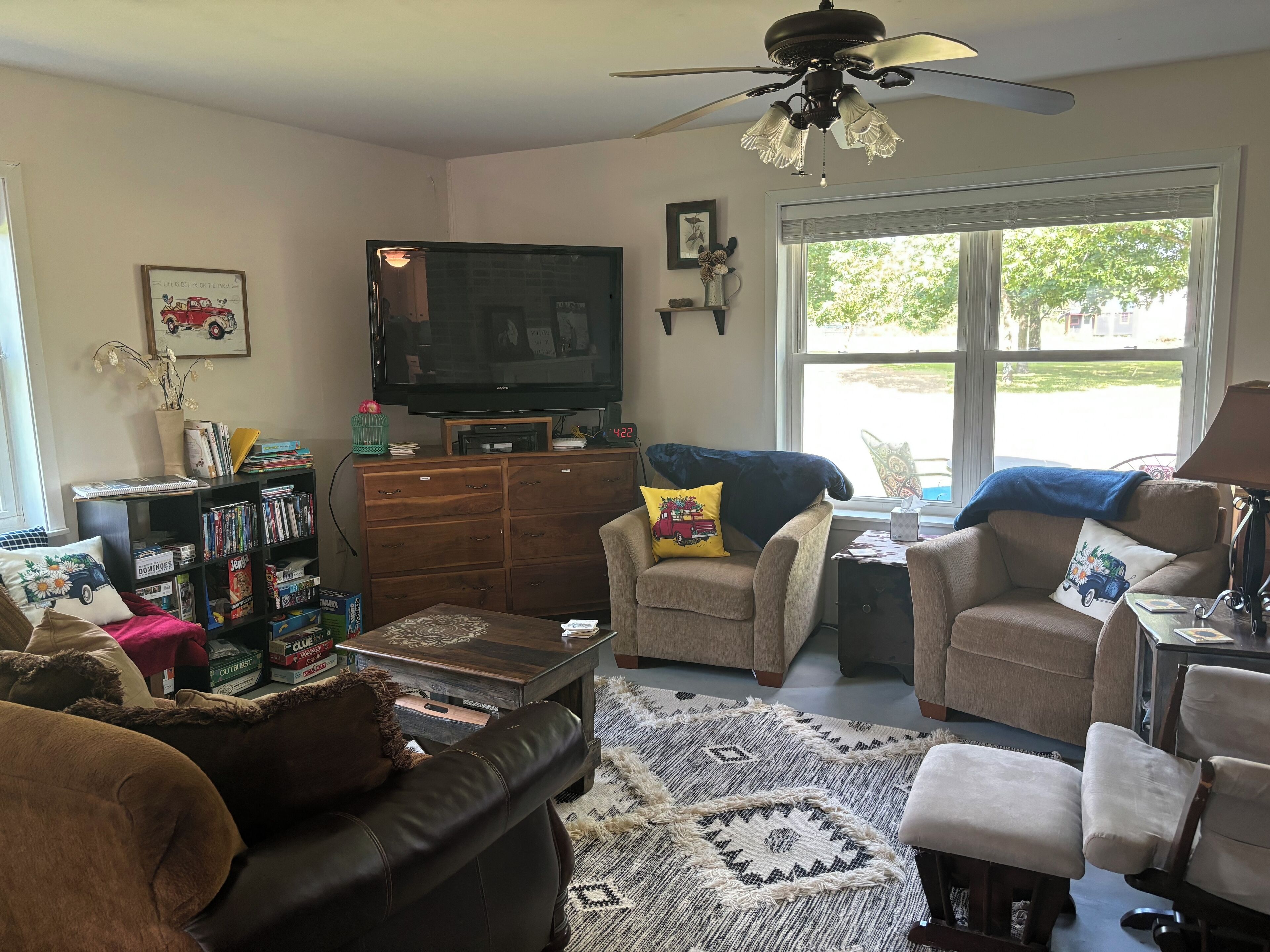 A living room with a television, a ceiling fan, a bookshelf, and a window with a view of the outdoors.