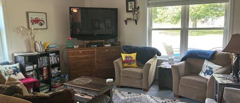 A living room with a television, a ceiling fan, a bookshelf, and a window with a view of the outdoors.