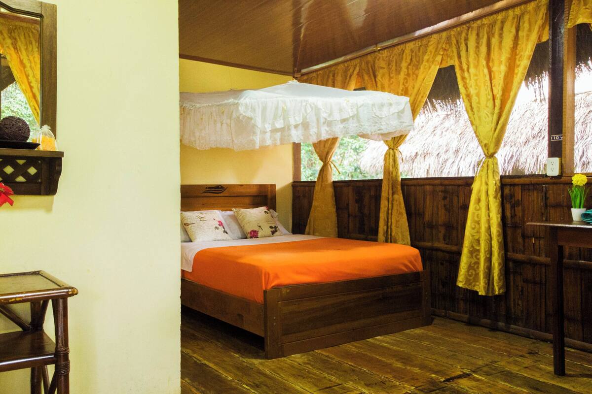 A wooden bed with an orange bedspread, a bedside table with a vase of flowers, and a window with yellow curtains.