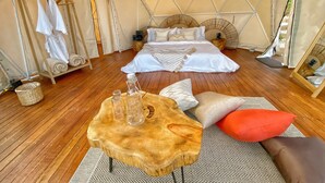 Une chambre aménagée sous une tente, comprenant un lit, une table en bois avec des bouteilles, un porte-vêtements suspendu et une petite table d’appoint.